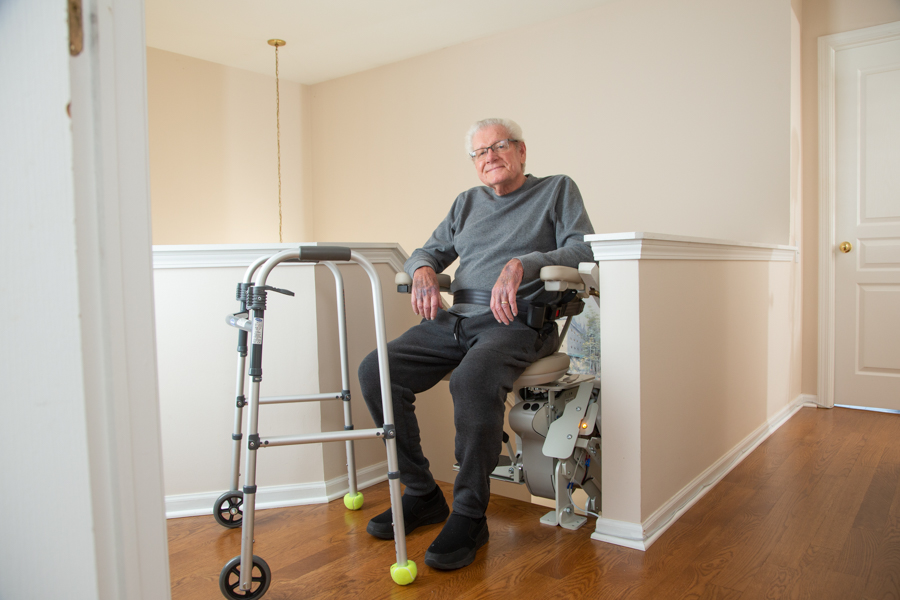 senior sitting on stairlift at top landing of stairs with walker in front of him on the landing